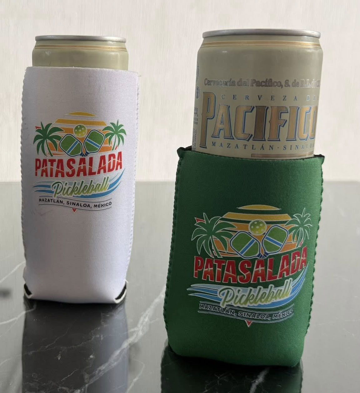 Two tall beer cans in Pata Salada Pickleball branded cozies—one white and one forest green—sitting on a reflective surface. The green cozy is holding a Pacifico beer can.