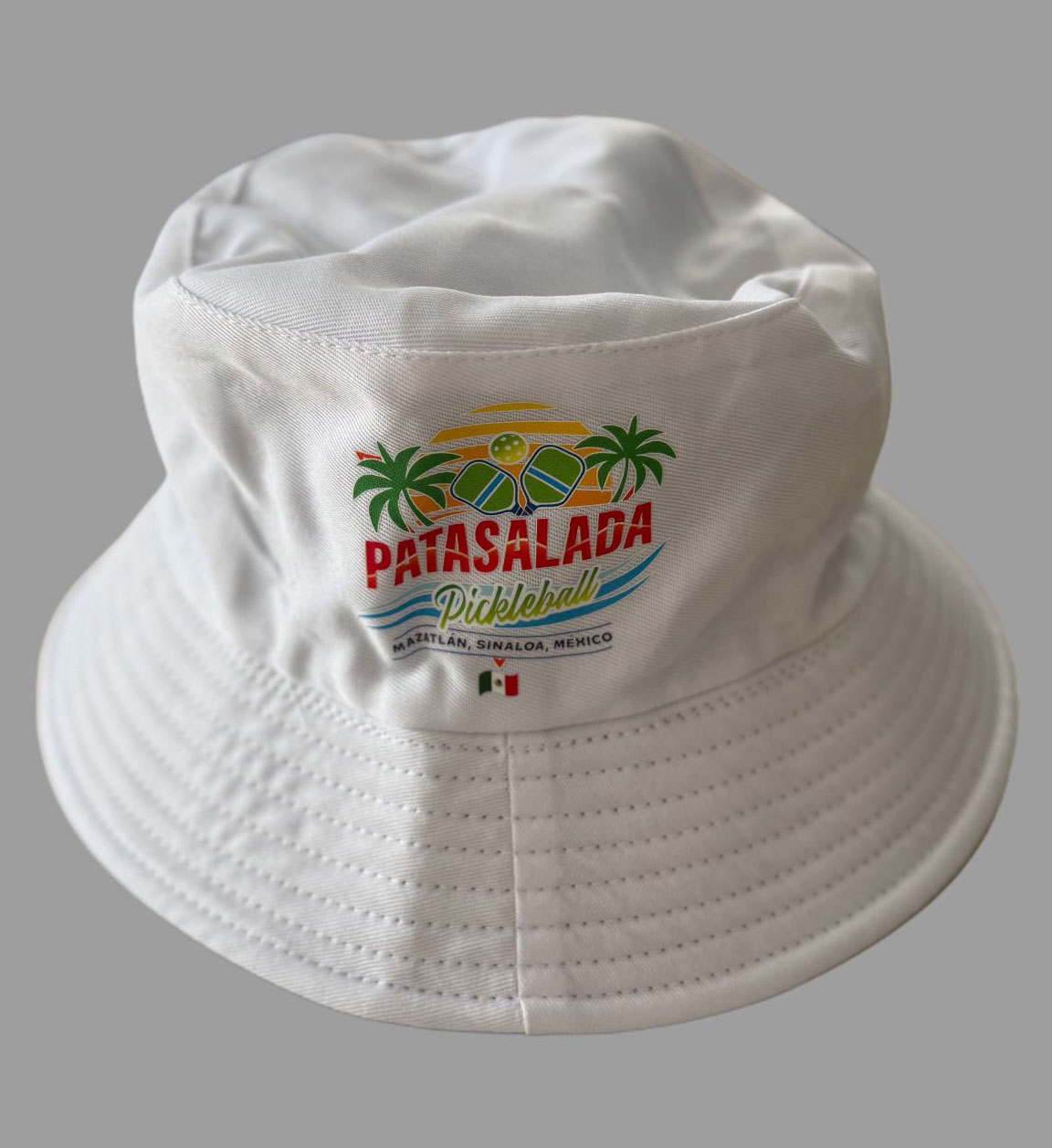 A white cotton bucket hat featuring the Pata Salada Pickleball logo with palm trees, a sunset, and a small Mexican flag centered below the text.