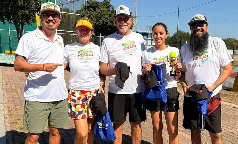 Five pickleball players in Pata Salada branded shirts smiling and holding a trophy and tournament gift bags at an outdoor court in Mazatlán.